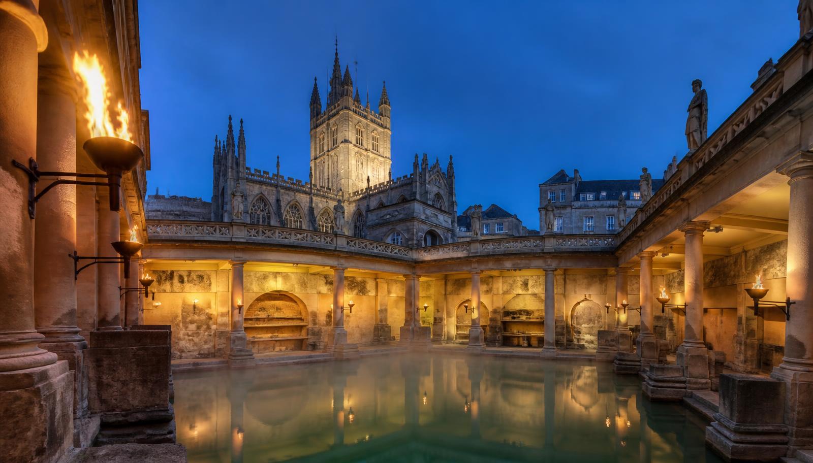 Los Roman Baths de Bath iluminados al anochecer con la torre de Bath Abbey al fondo